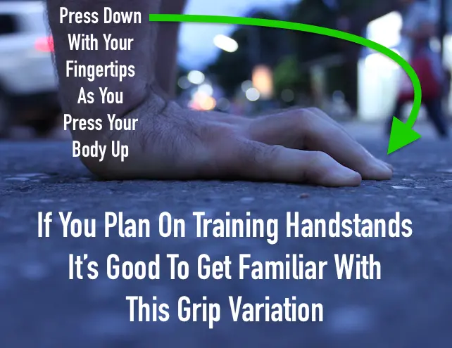 A close up image of just the hands on a cement road, showing the fingertips curled and pressing into the ground. This hand position is a good pre-requisite for eventually training handstands